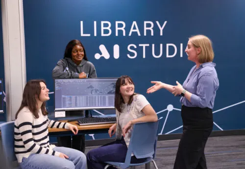 Three female students engaging in conversation with a female professor in front of a navy blue wall featuring "Library AI Studies"