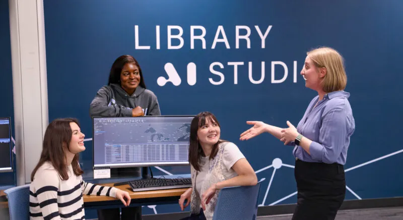 Three female students engaging in conversation with a female professor in front of a navy blue wall featuring "Library AI Studies"