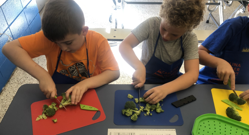 Two children using a knife and cutting board to cut broccoli.