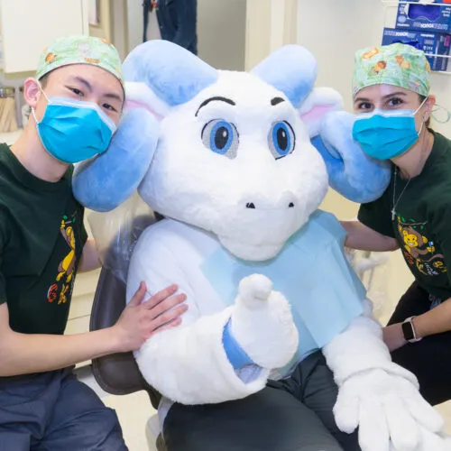 Rameses relaxes in a dentist's chair next to two dental students giving a thumbs up