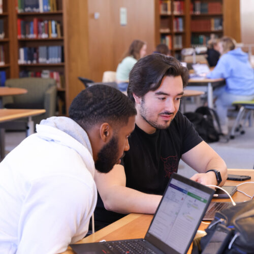 Two male students sit at a table inside Davis Library looking at their laptops.