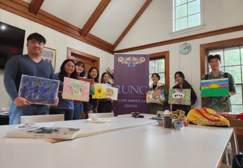A small group of individuals standing next to a UNC Asian American Center sign holding artwork and posing for a picture.