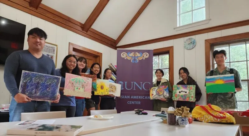 A small group of individuals standing next to a UNC Asian American Center sign holding artwork and posing for a picture.