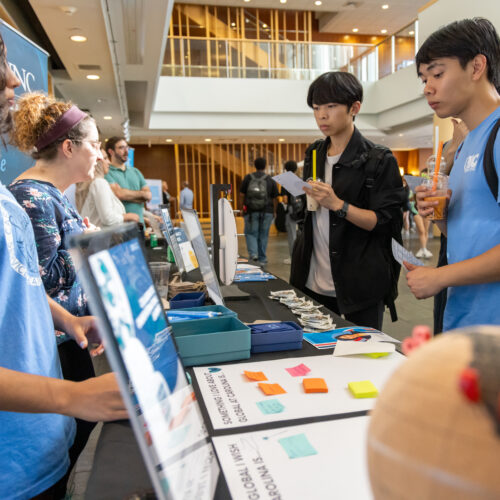 Two male students holding drinks stop at a table containing information about UNC Global Affairs programs.