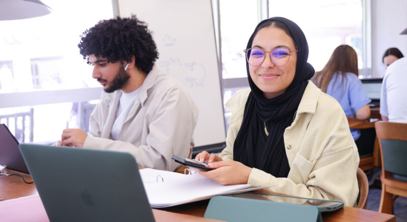 Female student in library working on her computer while smiling at the camera.