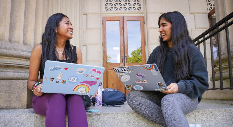 Two students working with laptops on the stairs outside of Wilson Library. The students are talking to each other. The wooden entrance door can be seen in the background.