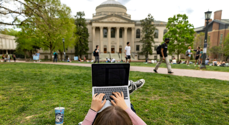 Student laying in the grass in front of Polk Place typing on laptop.