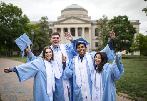 A group of Carolina graduates in cap and gown standing in front of Wilson Library.