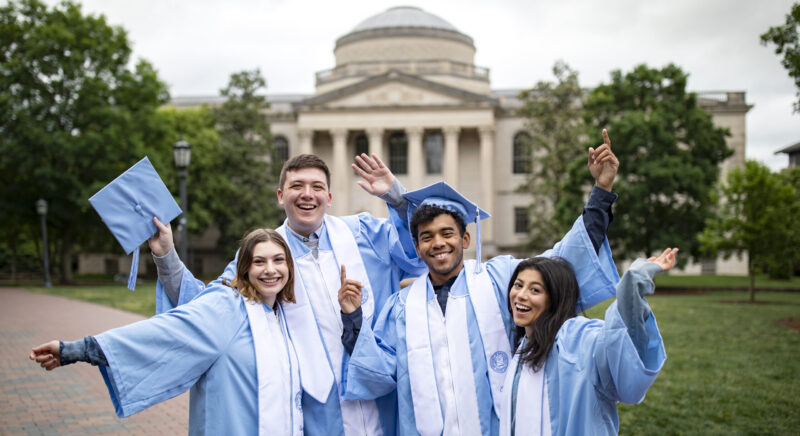 A group of Carolina graduates in cap and gown standing in front of Wilson Library.