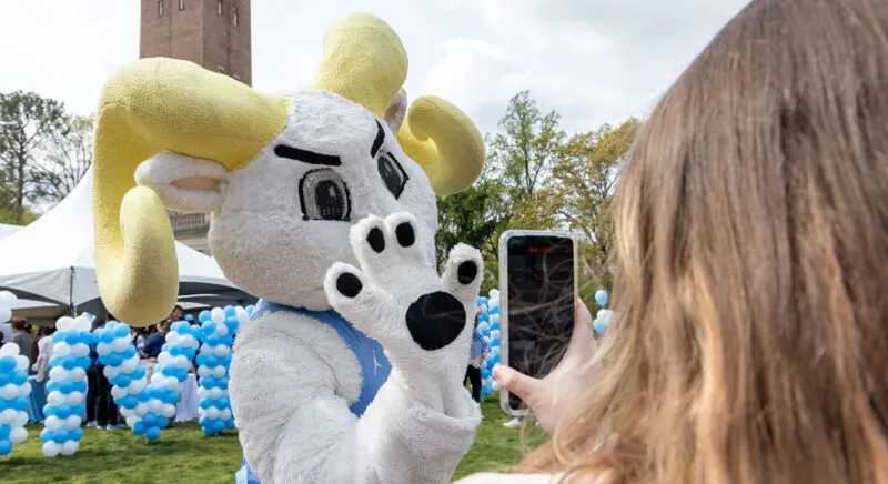 Rameses waving at a student taking a photo on their phone, in front of the Bell Tower on GiveUNC