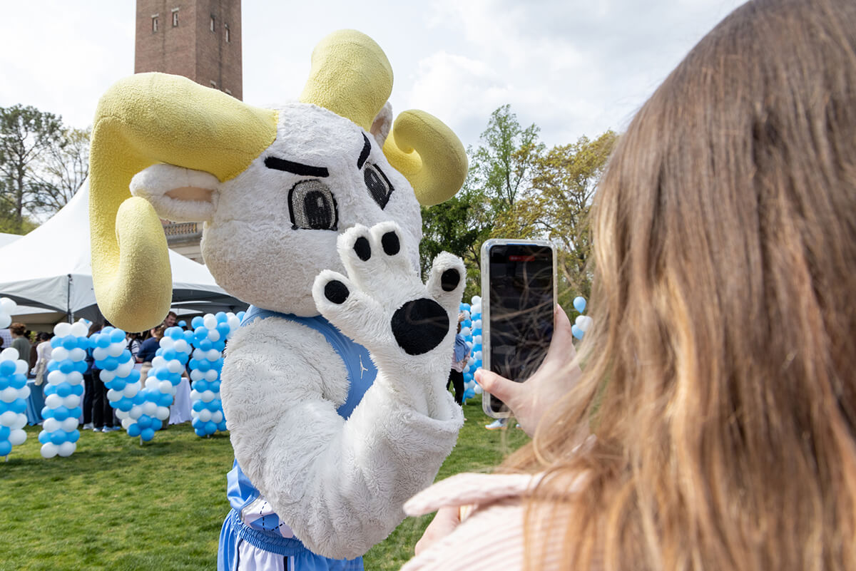 Rameses waving at a student taking a photo on their phone, in front of the Bell Tower on GiveUNC